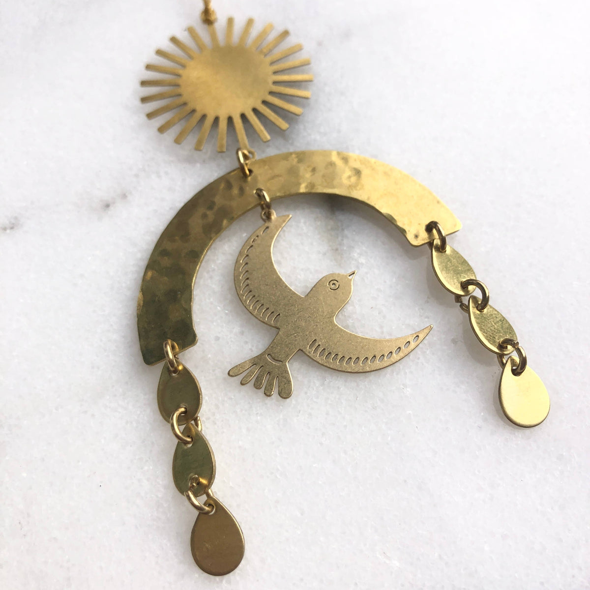 A handmade brass dangle ornament featuring a sunburst, a hammered crescent with a bird silhouette, and teardrop-shaped charms, displayed on a white marble surface.