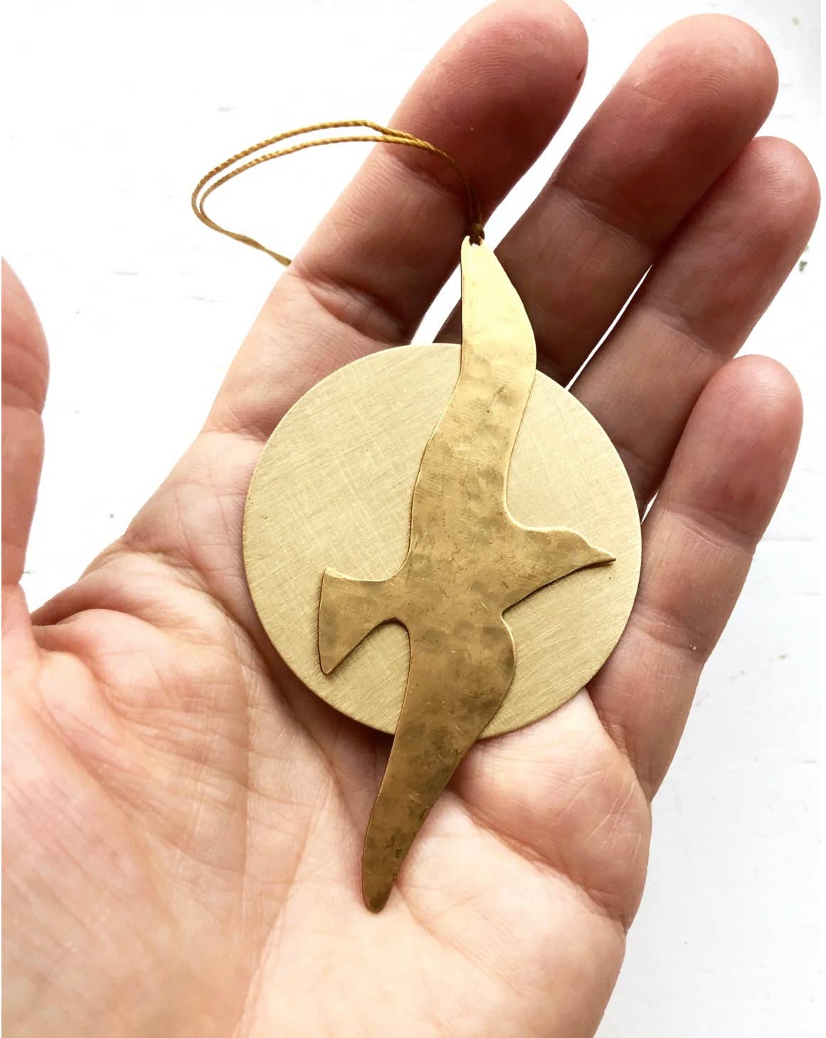 A hand holding a hammered brass ornament featuring a dove silhouette over a circular background, with a thin hanging string.