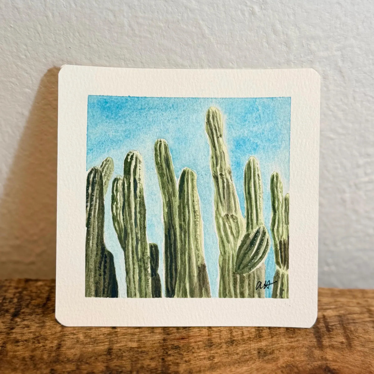 A square watercolor art print depicting green saguaro cacti against a bright blue sky, mounted on a white card and signed by Tiny Desert Studio artist in the right hand corner.