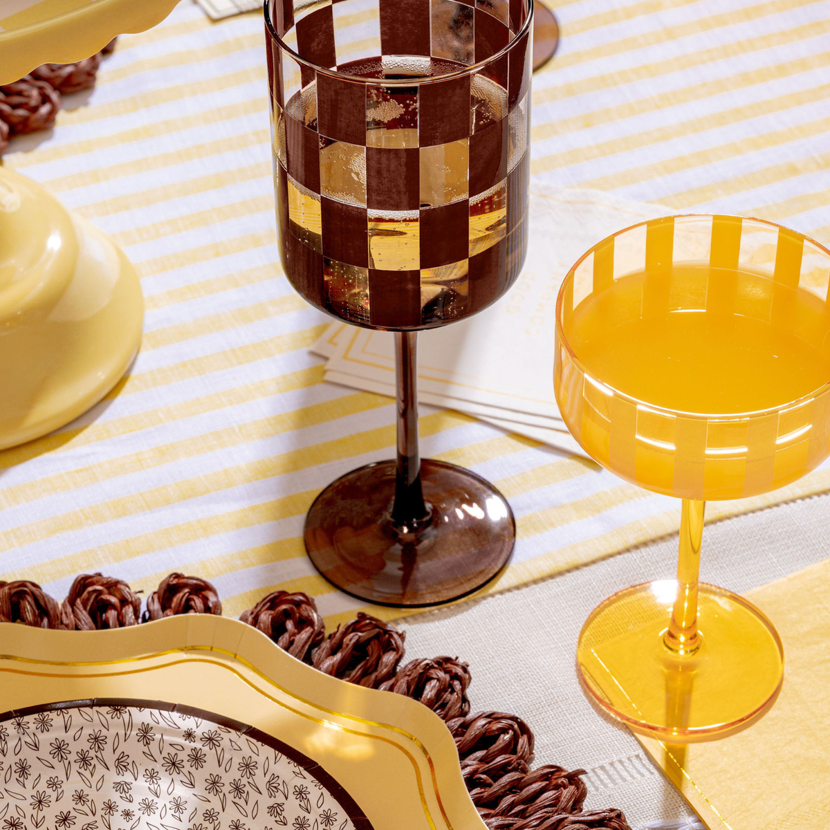 A styled tabletop scene with two decorative drinking glasses on a striped tablecloth. The taller glass has a dark brown and amber checkerboard pattern, while the shorter coupe-style glass is translucent amber yellow.