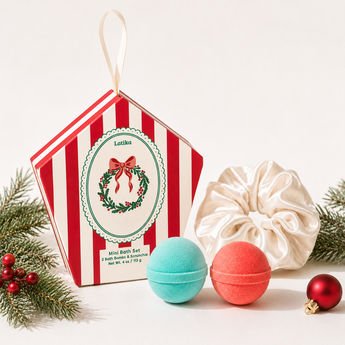 A Christmas-themed bath set gift box by Lotika, shown with a cream satin scrunchie and two bath bombs in mint green and coral red, styled with pine branches and a red ornament on a white background.