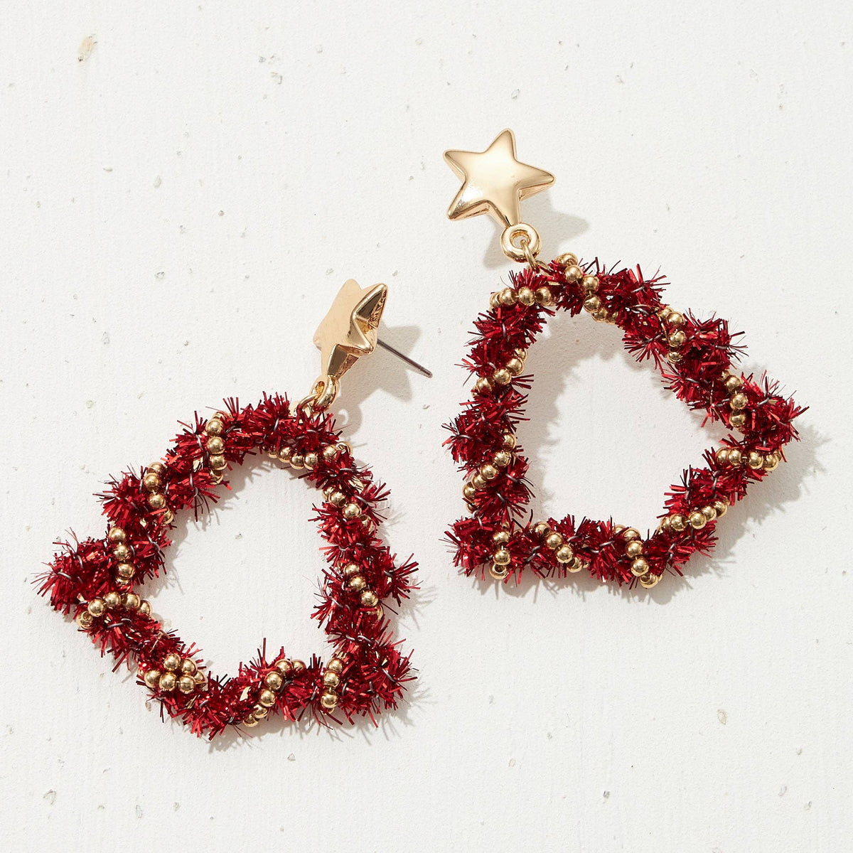 A pair of gold-tone dangle earrings with star-shaped posts and triangular hoops wrapped in textured red tinsel with small gold beads, displayed on a light off-white background.