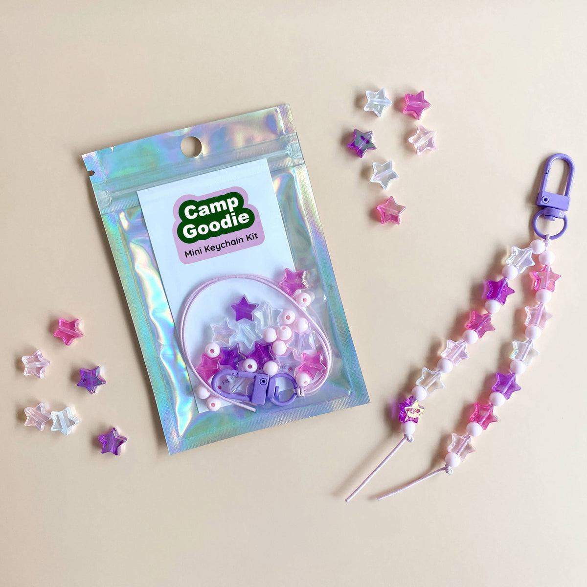 A Camp Goodie DIY Keychain Kit shown with its iridescent packaging and a completed lavender and white star-patterned keychain laid out on a beige background with loose star embellishments.