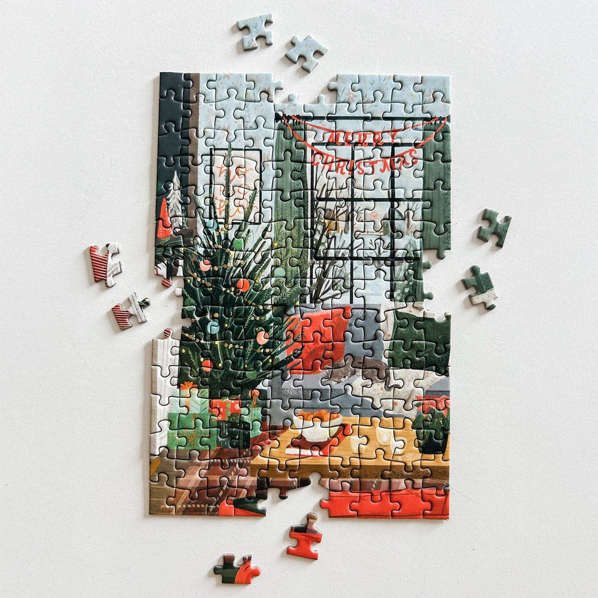 A partially assembled jigsaw puzzle showing a cozy indoor holiday scene with a Christmas tree, warm window light, and red garlands, placed on a plain white surface with a few loose puzzle pieces scattered around.