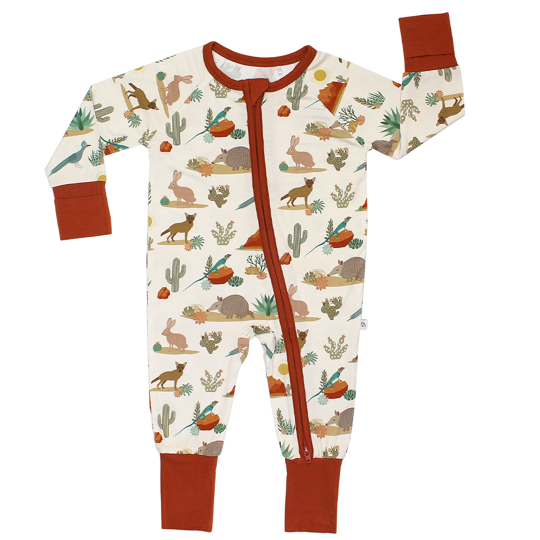 A baby long-sleeve footless sleeper with an all-over desert safari print on an off-white background, featuring rust-colored cuffs, collar, and zipper trim, and a matching print of cacti, rabbits, coyotes, and lizards. 