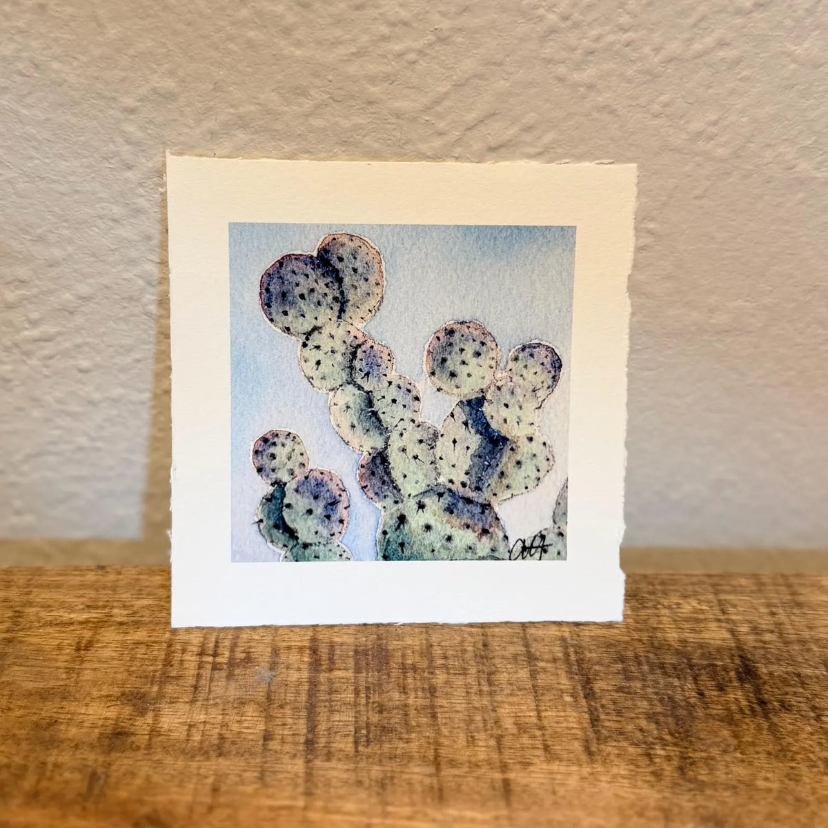 A square art print with a white border featuring a watercolor painting of a prickly pear cactus, hand painted and signed by artist in right hand corner. 
