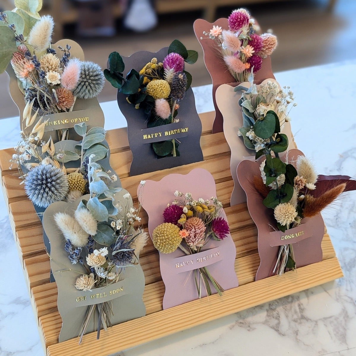 A wooden display stand holding six small dried flower bouquets in pastel-colored card sleeves, photographed on a marble-like surface with a softly blurred indoor background.