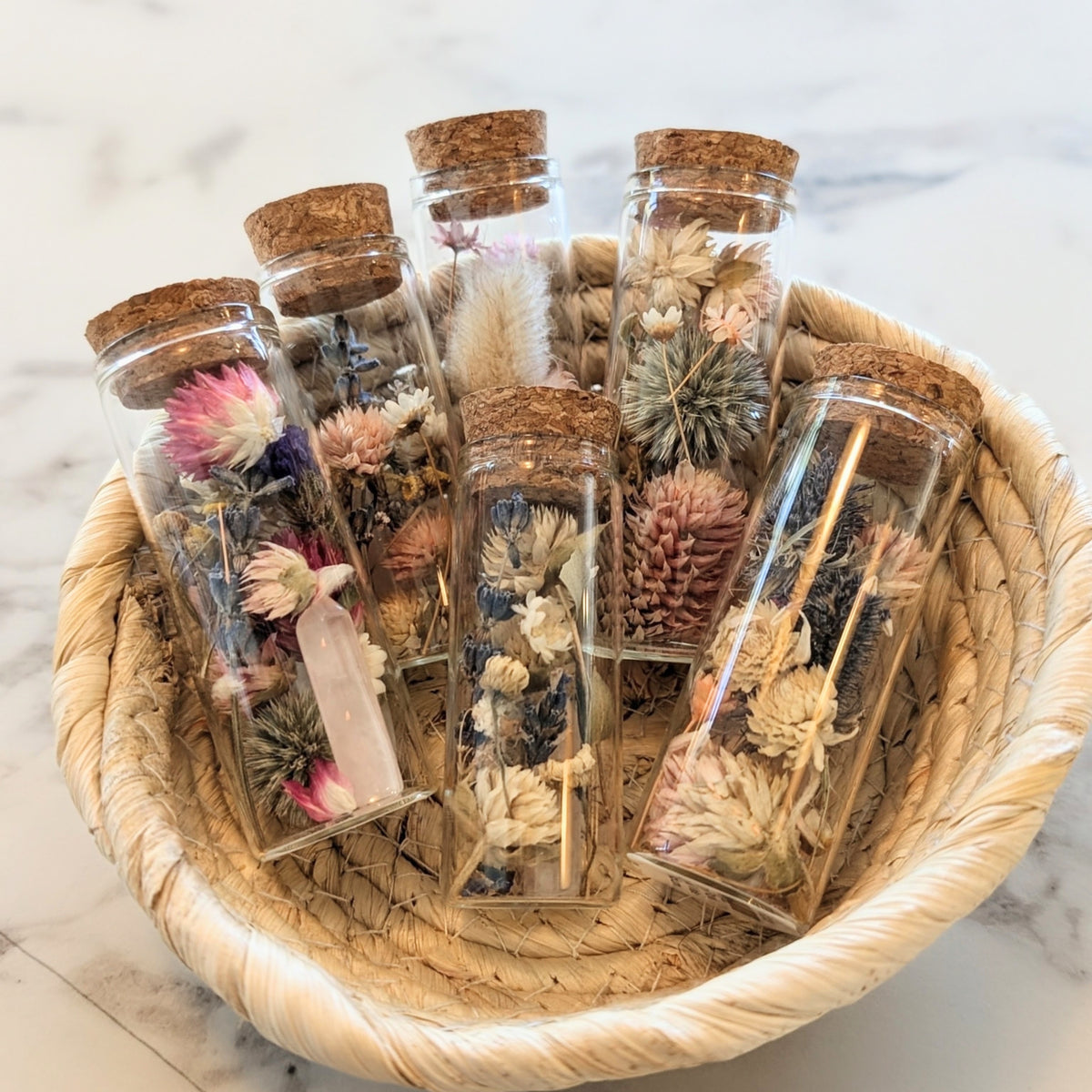 Six small clear glass vials with cork stoppers, each containing dried botanicals and crystals.