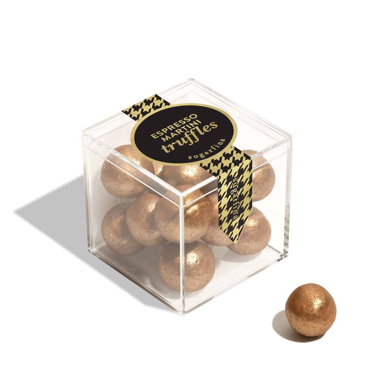 A clear acrylic cube box filled with round metallic gold espresso martinis truffles, tied with a black and gold houndstooth ribbon and a round black label. A single truffle sits outside the box on a clean white background.