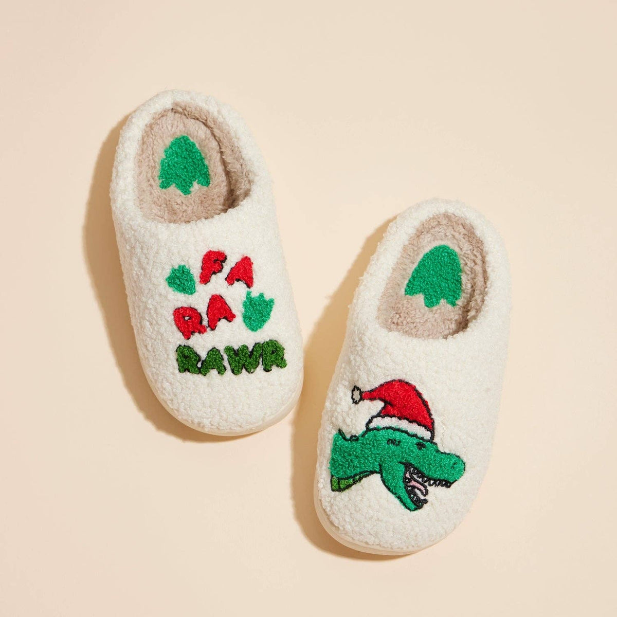 A pair of cream sherpa house slippers with embroidered holiday designs, one featuring FA RA RAWR in red and green and the other featuring a green alligator wearing a red Santa hat, set against a solid beige background.