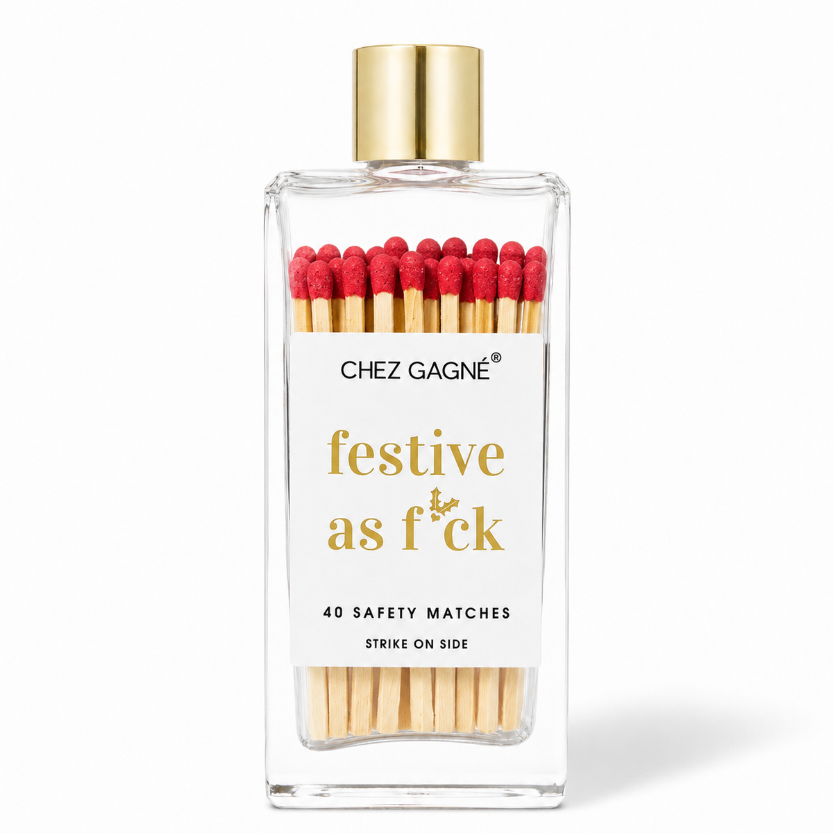 A clear glass bottle of Chez Gagnée safety matches with a gold cap and a white label reading 'festive as f*ck'. The matches inside have red tips and light wood-colored bodies, visible through the transparent bottle against a white background.