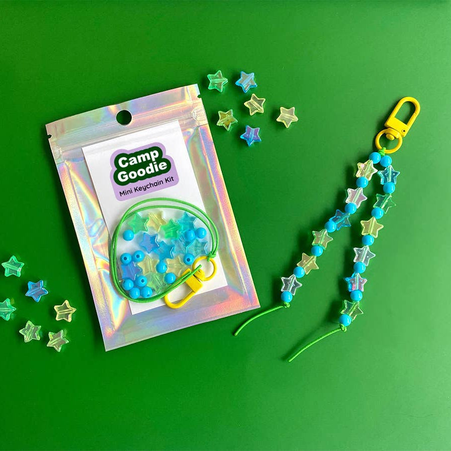 A Camp Goodie Mini Keychain Kit shown with its holographic packaging on a bright green background, alongside a completed keychain featuring blue and green star beads, a yellow clasp, and a green cord.
