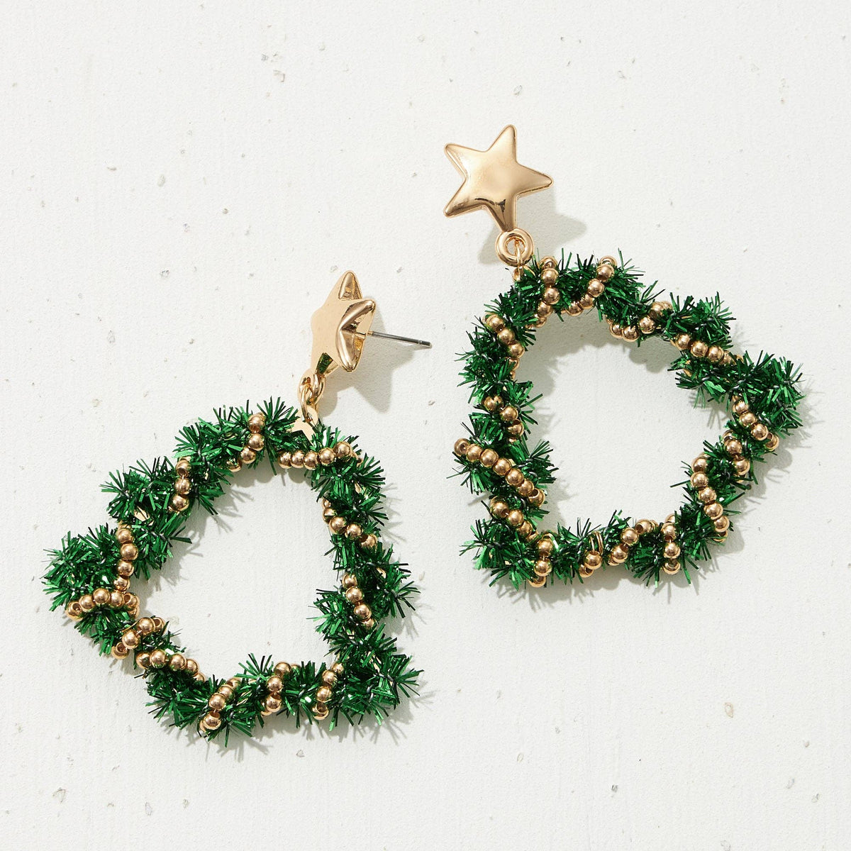 A pair of gold-tone and green tinsel drop earrings shaped like Christmas wreaths with small gold beads and star-shaped studs, displayed on a light textured background.