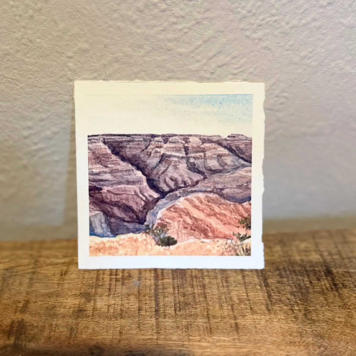 A small square watercolor art print depicting the grand canyon landscape with layered red, brown, and gray rock formations and a pale blue sky, signed by tiny desert studio in right hand corner.