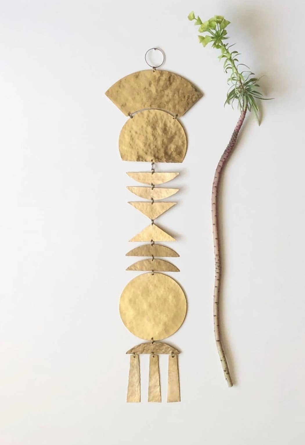 A hammered gold metal wall hanging with geometric shapes, displayed vertically next to a thin brown branch with small green leaves against a plain white background.