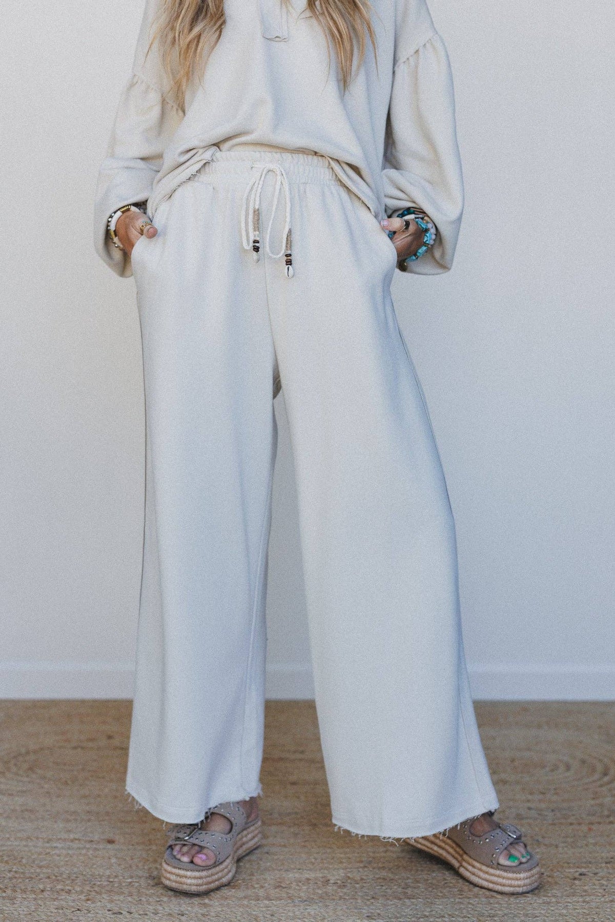 A person wearing a light cream or off-white lounge pant consisting of wide-leg drawstring pants, paired with espadrille sandals, standing against a plain white wall.