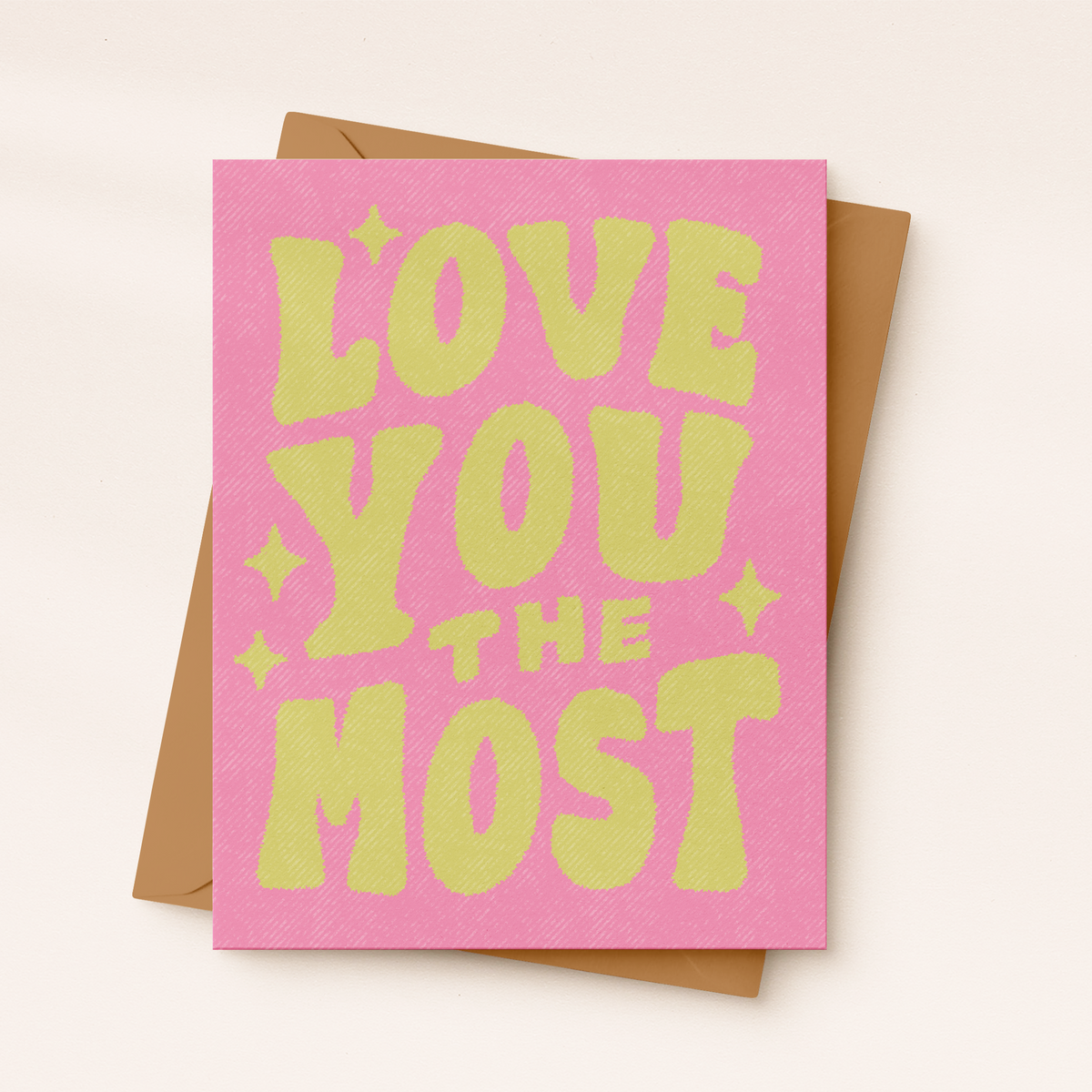 A pink greeting card with bold yellow-green retro text reading 'LOVE YOU THE MOST' and a brown envelope behind it, photographed on a light neutral background.