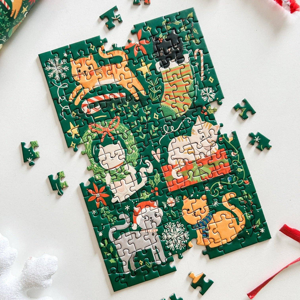 Three partially assembled jigsaw puzzle pieces featuring festive Christmas-themed cat illustrations on a dark green background, arranged on a white surface with a few loose puzzle pieces around them.
