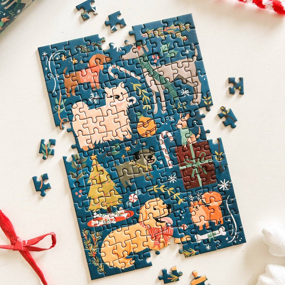 A colorful dog-themed jigsaw puzzle, partially assembled and photographed from above on a white surface with a few loose pieces scattered around. The puzzle features illustrated dogs, a Christmas tree, candy canes, and holiday decorations on a dark blue background.