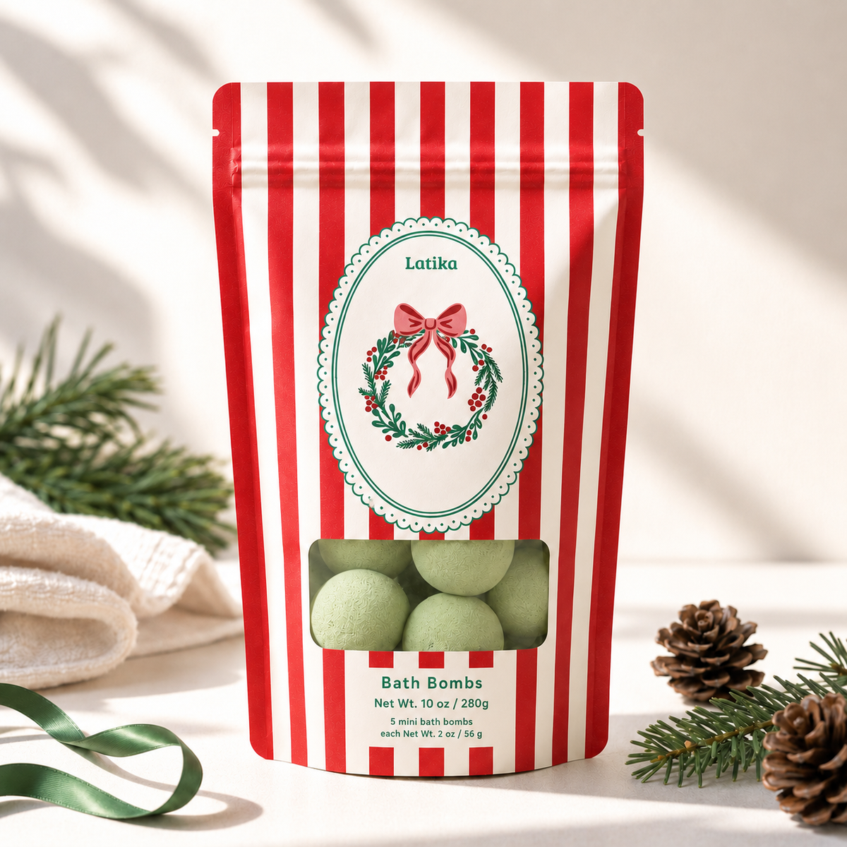 A red and white striped stand-up pouch of Lotiko holiday-themed bath bombs, displayed on a light surface with pine branches, a green ribbon, and a textured towel nearby.