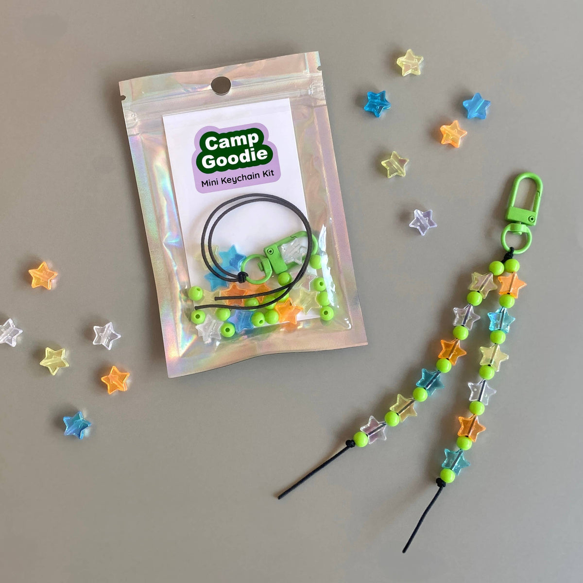 A Camp Goodie mini keychain kit shown with its iridescent zip pouch packaging and a completed example keychain laid out on a neutral gray background, surrounded by loose star-shaped beads.