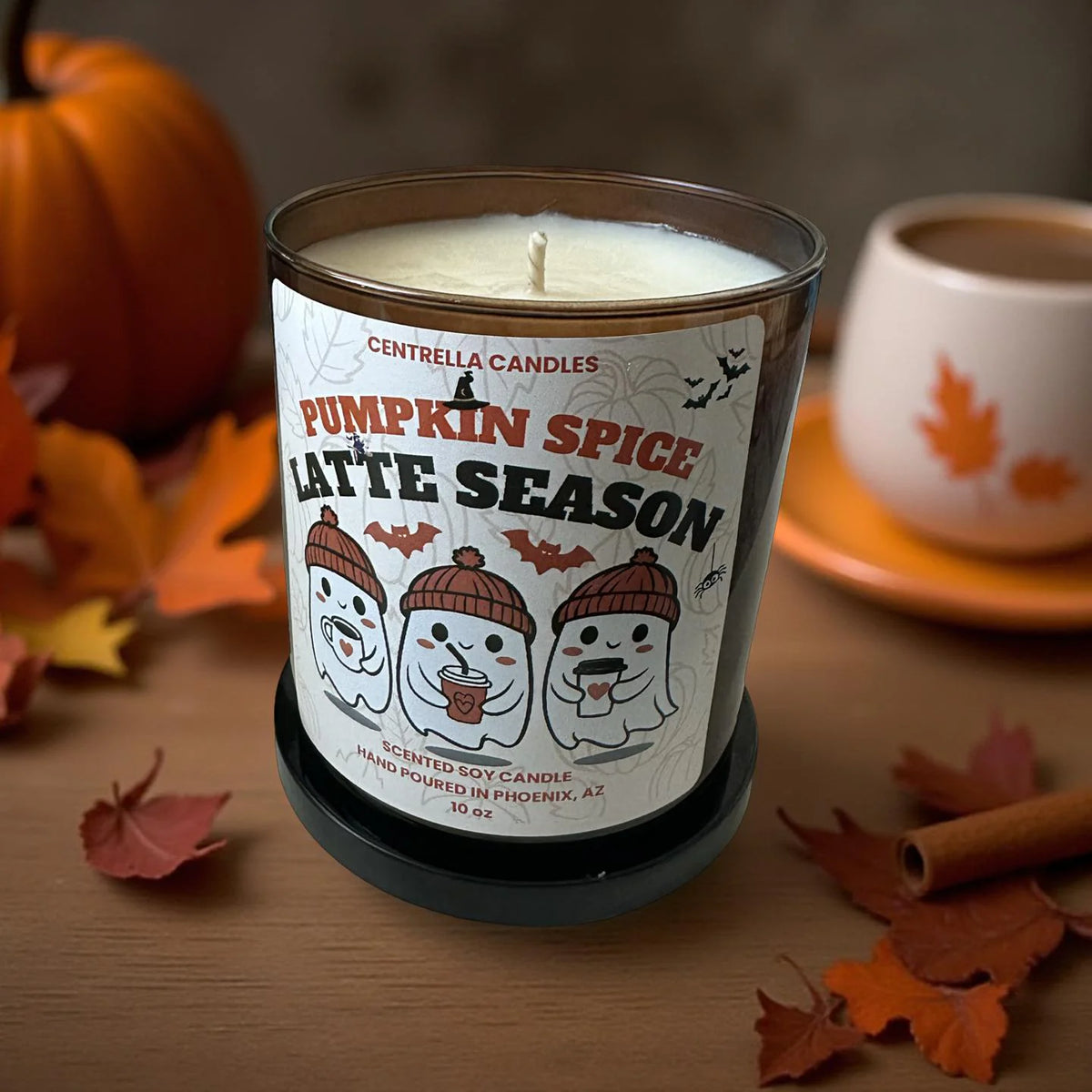 A 16 oz scented soy candle by Centrella Candles in a clear glass jar with a black base, labeled 'Pumpkin Spice Latte Season'. The candle is styled on a wooden table with autumn leaves, a pumpkin, and a warm mug in the background.