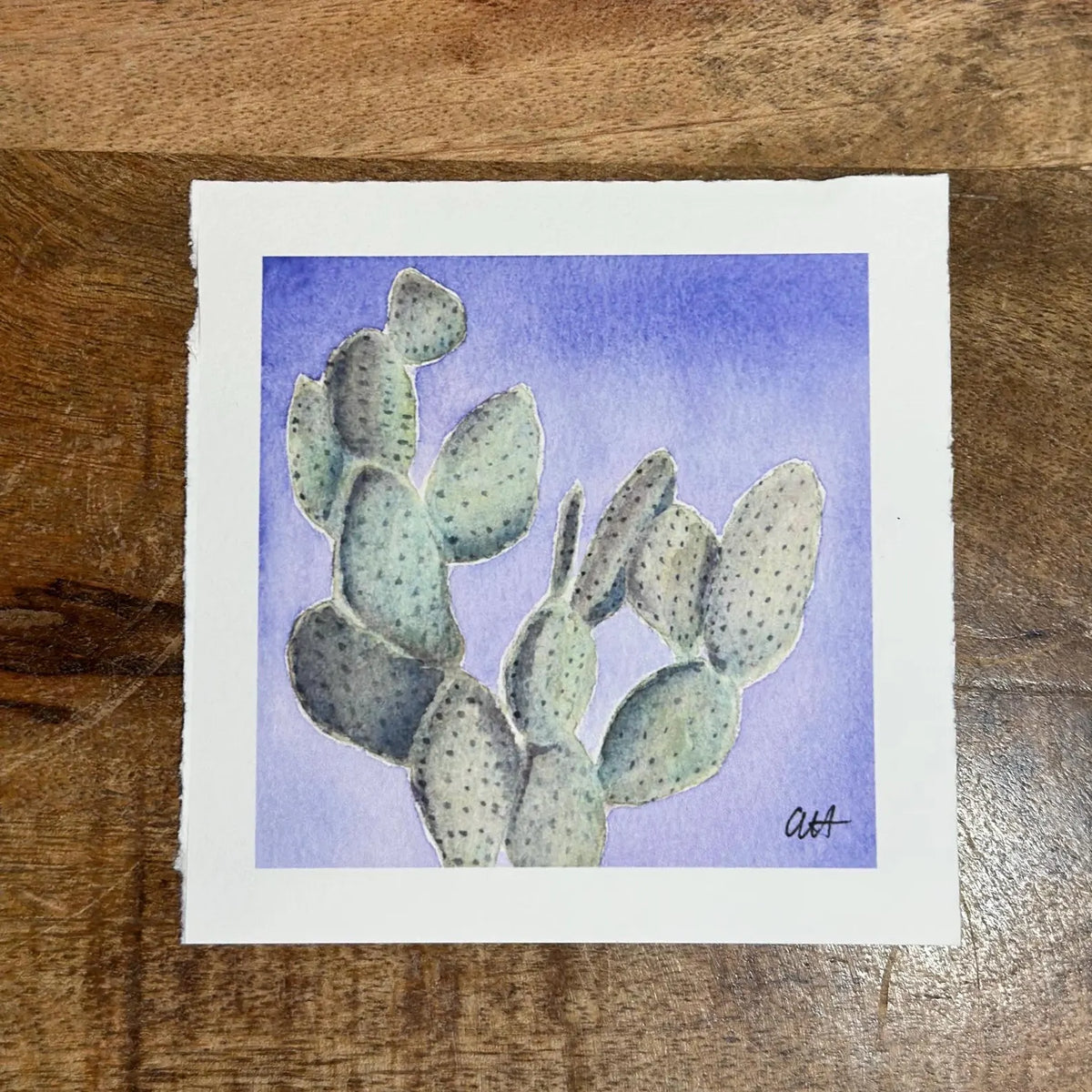 A square watercolor painting of a prickly pear cactus on textured white paper, placed on a wooden surface. The painting features light green cactus pads with small dark dots against a soft blue-violet watercolor background, signed by artist in the bottom right corner.