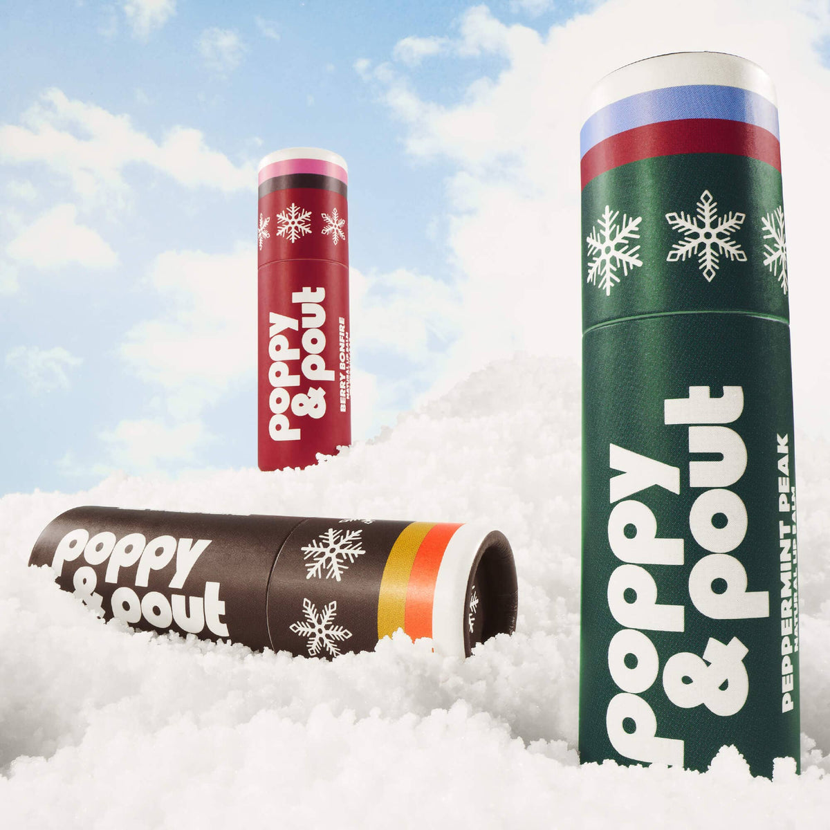 Three cylindrical Poppy & Pout lip balm tubes standing in white faux snow against a blue sky with clouds. One tube is red, one is dark green labeled Peppermint Peak, and one is dark brown.