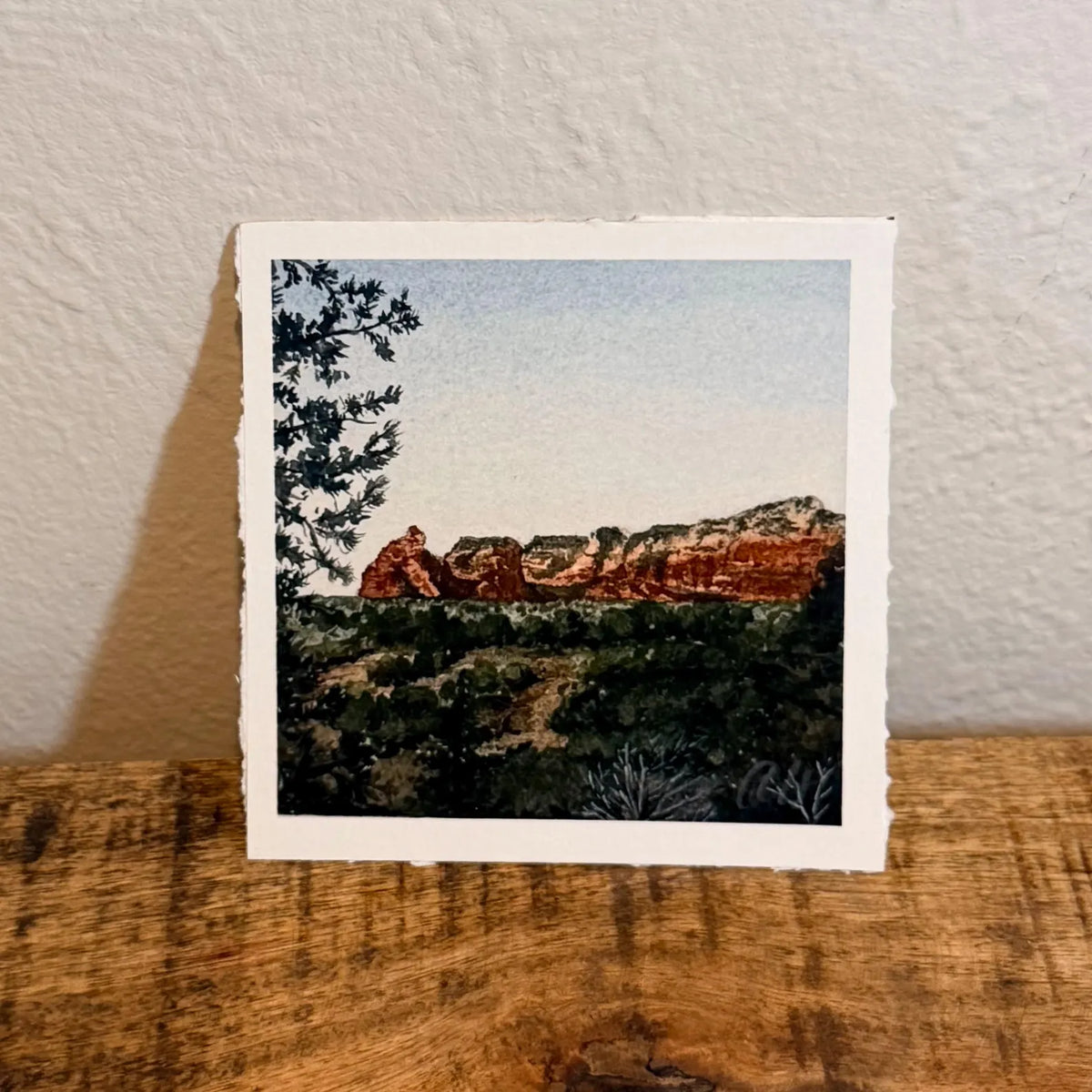 A square watercolor art print depicting a desert landscape with red rock formations, green shrubs, and a pale sky, inspired by Sedona by Tiny Desert Studio 