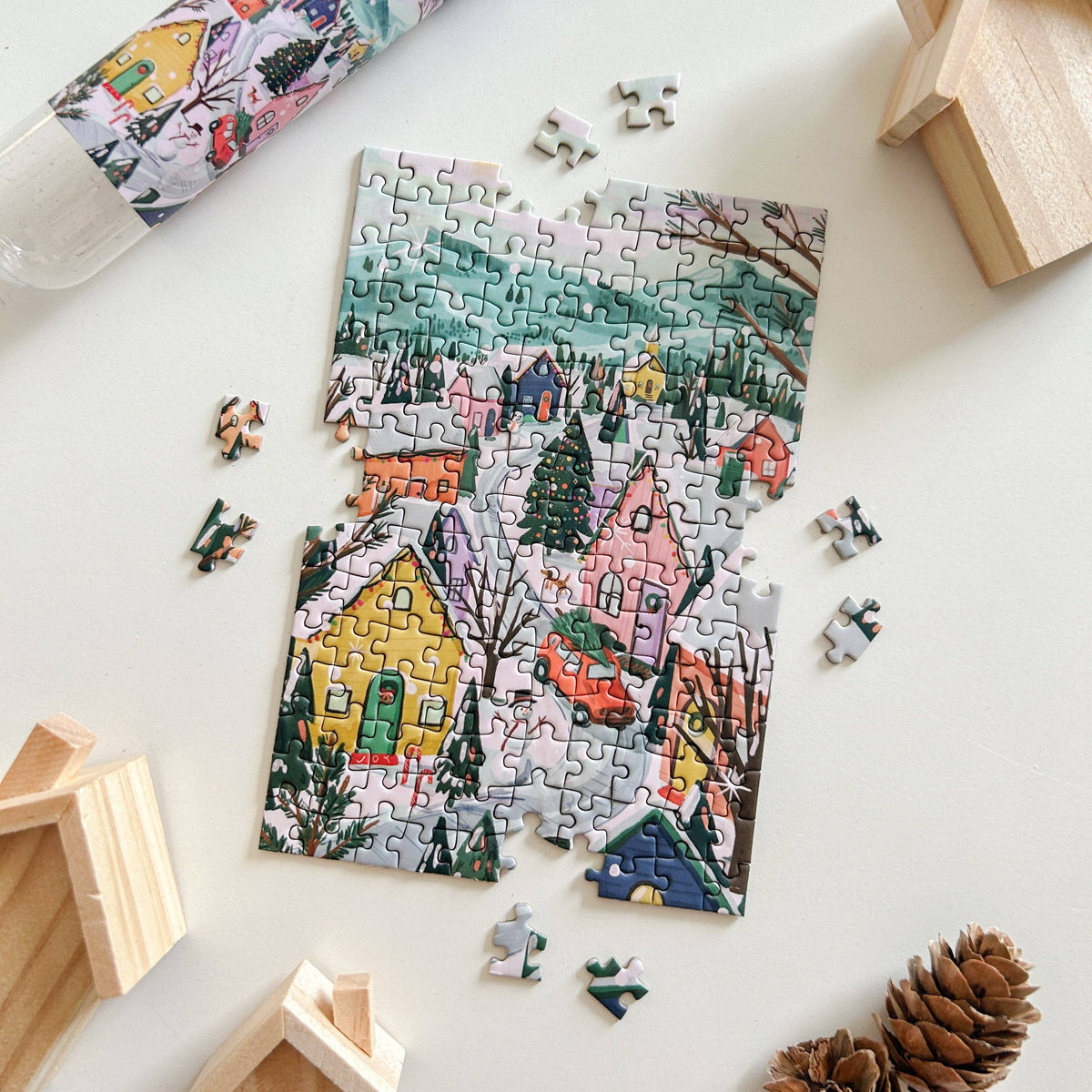 A partially completed jigsaw puzzle showing a colorful winter village scene with houses, trees, and snow, arranged on a light surface with a few loose puzzle pieces around it. A rolled tube of puzzle pieces and wooden house-shaped props are visible in the background.