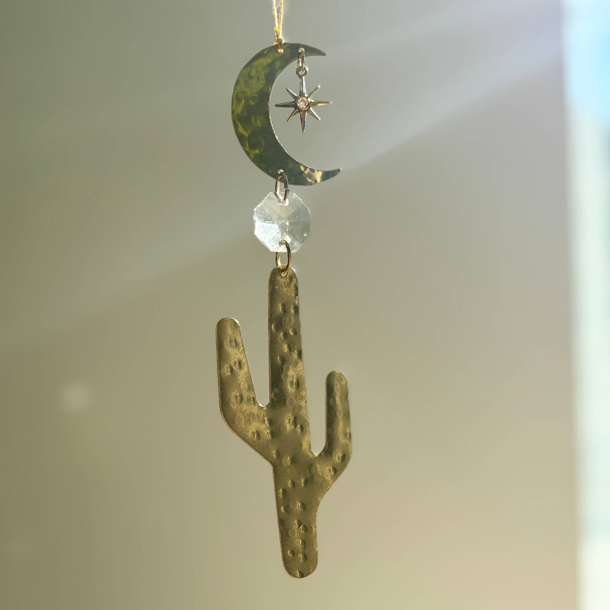 A hanging decorative ornament suncatcher featuring a hammered gold-toned cactus, a gold crescent moon with a small star detail, and a faceted clear bead, suspended against a softly lit neutral background.