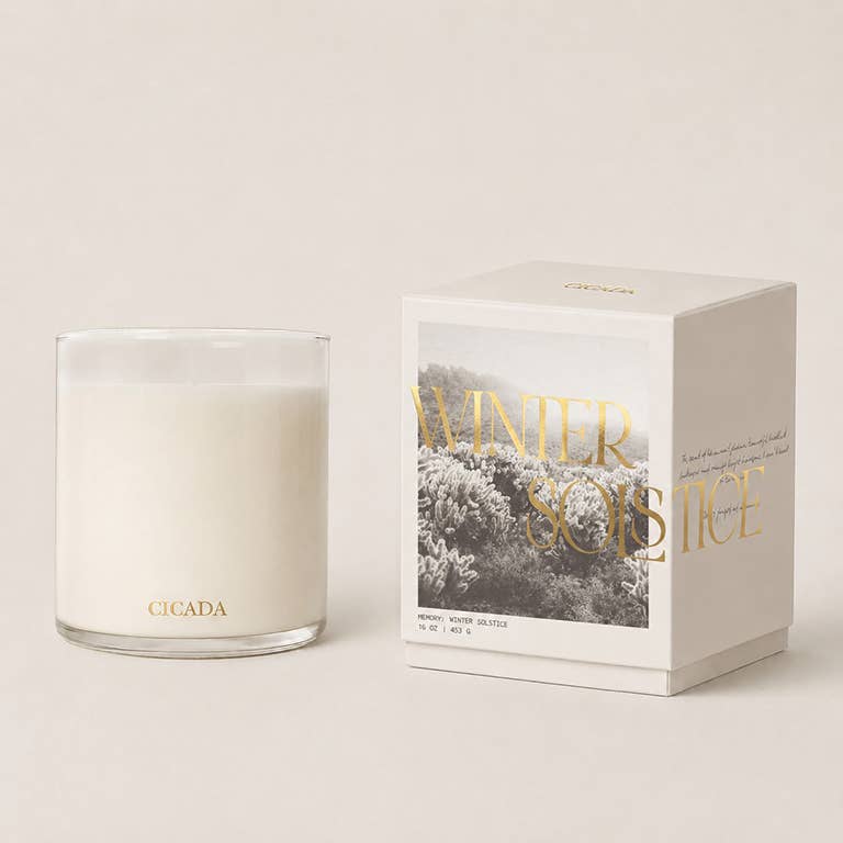 Winter Solstice Candle, in a glass Cicada branded Glass votive, next to the box it comes in , a black and white box labeled with Winter Solstice in Gold. 