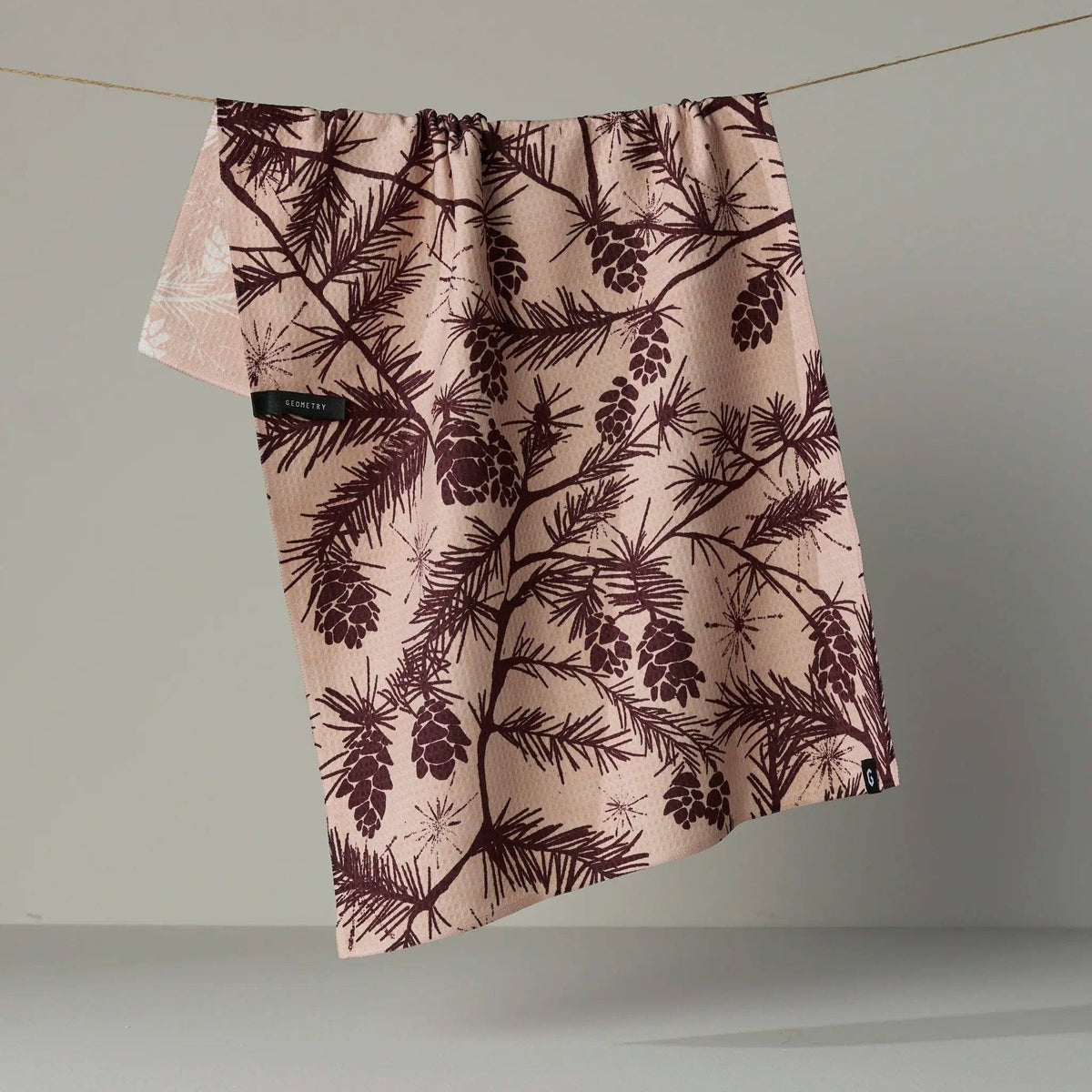 A fabric towel or cloth with a dark plum-brown pine branch and pinecone pattern on a dusty pink background, hanging from a thin wire against a neutral gray studio backdrop.