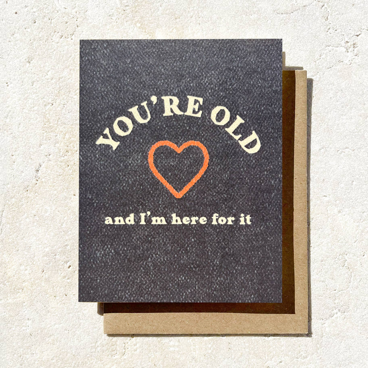 Greeting card with text 'You're old and I'm here for it' and a heart on a textured surface.