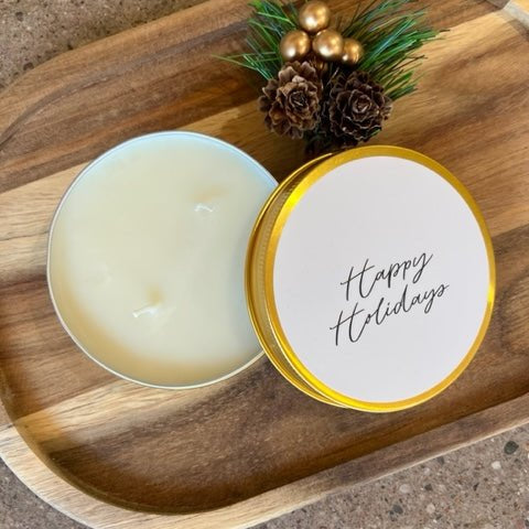 An open candle tin with creamy off-white wax sits beside its lid on a wooden tray. The lid has a white label with 'Happy Holidays' in black script and a gold rim.