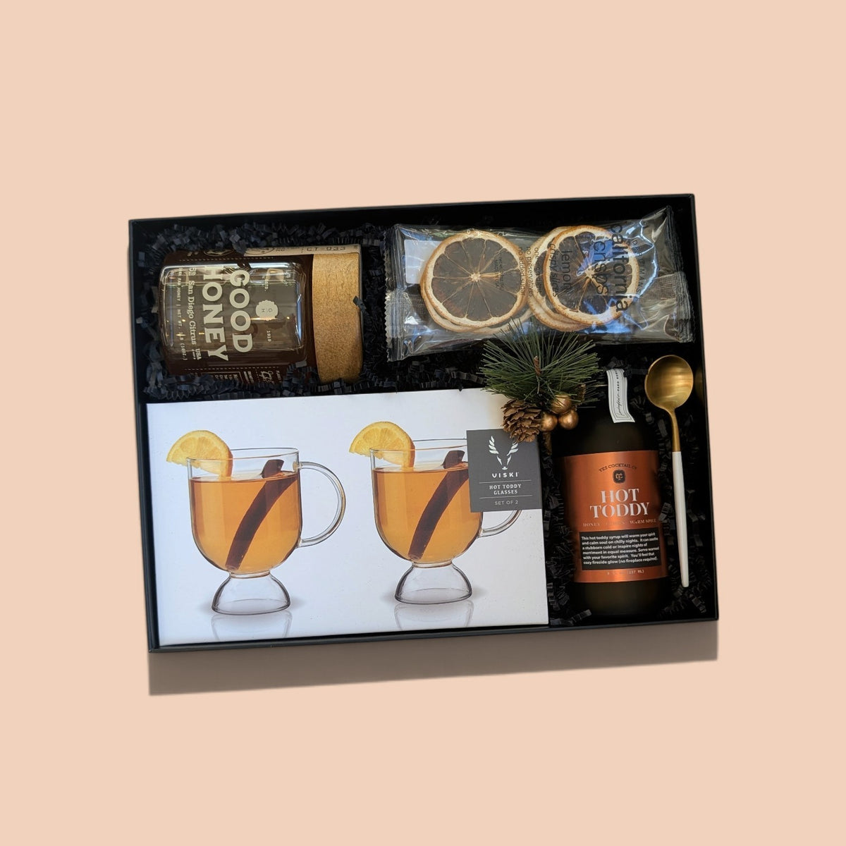 A hot drink gift set in a black presentation box, featuring a bottle of hot toddy, honey, dried orange slices, a small pine sprig, a gold spoon, and printed mugs with cinnamon and lemon illustrations.
